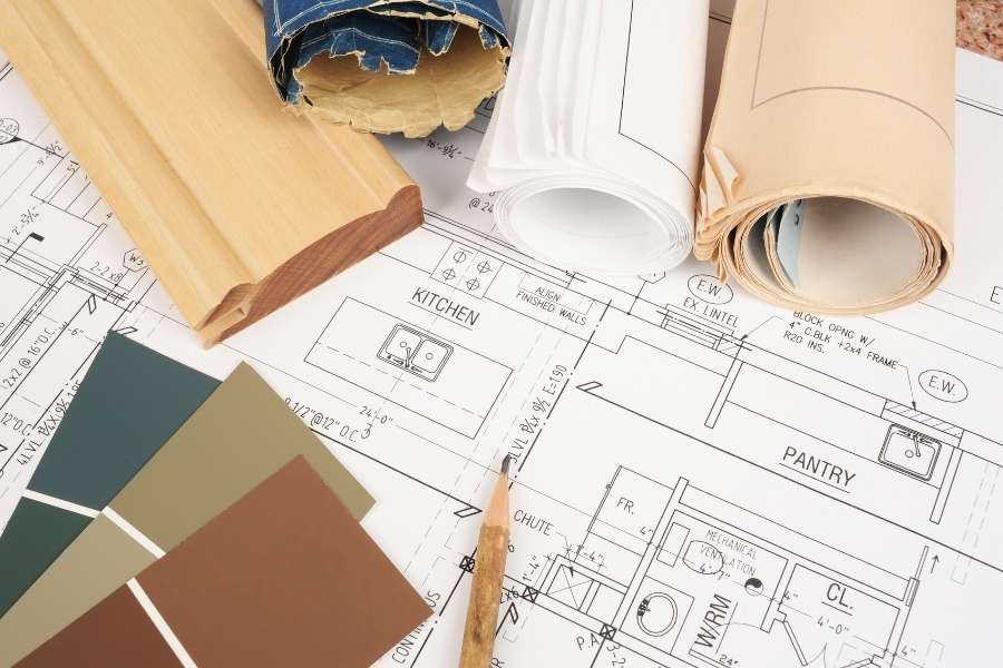 Des plans architecturaux, des rouleaux de papier et des échantillons de peinture posés sur une table pour préparer une rénovation de A à Z.