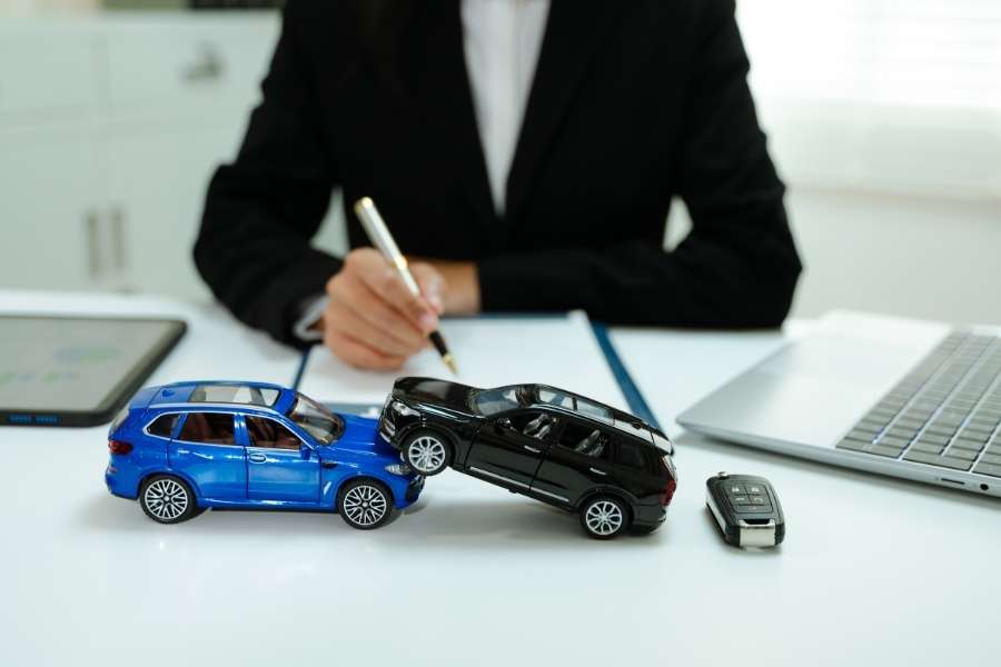 Une personne en costume prépare un dossier avec deux petites voitures miniatures accidentées pour illustrer la prime d'assurance auto.