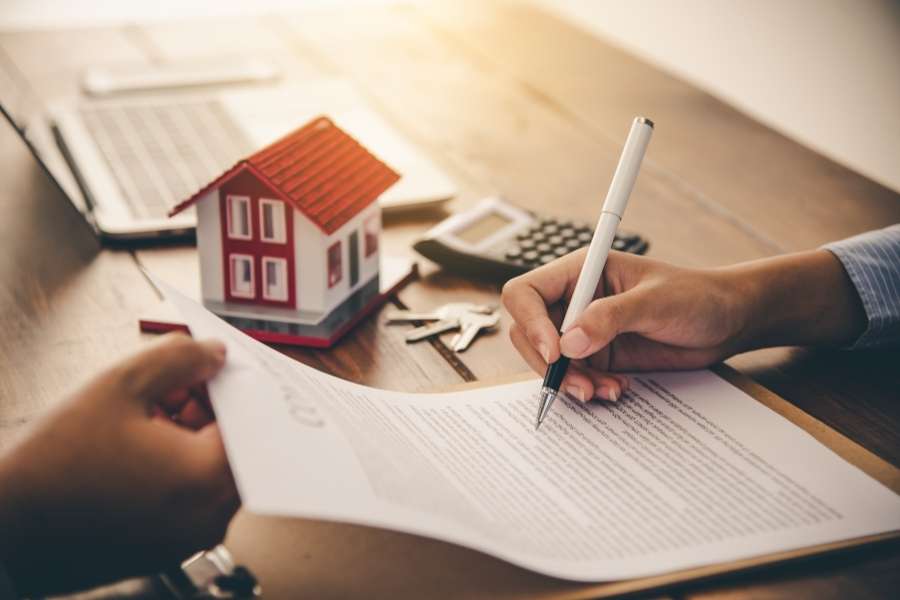 Une personne signe un contrat de prêt sur un bureau en bois avec une maquette de maison et une calculatrice, représentant un crédit immobilier.