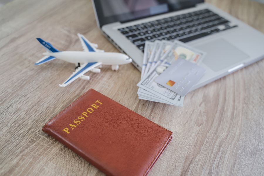 Un passeport en cuir marron, un avion miniature, des billets de banque et une carte bancaire posés sur un bureau en bois près d'un ordinateur portable.